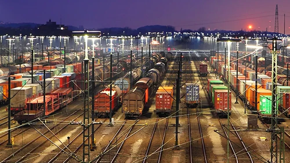 Recinto de una estación de ferrocarril con muchas vías, agujas y líneas eléctricas frente a la silueta de una ciudad.
