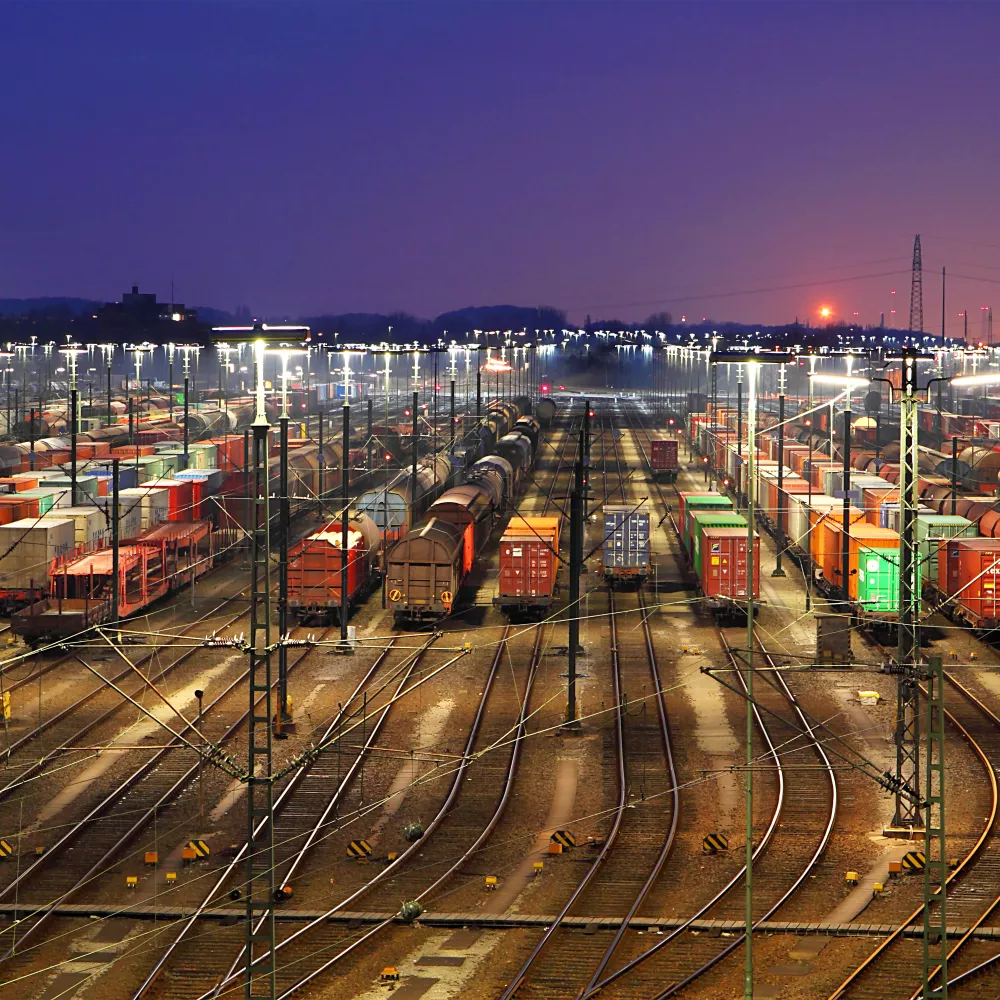 Numerosos trenes de carga y contenedores se alinean en un patio de maniobras iluminado al atardecer, con las vías del tren convergiendo en la distancia y el sol poniéndose en el horizonte.