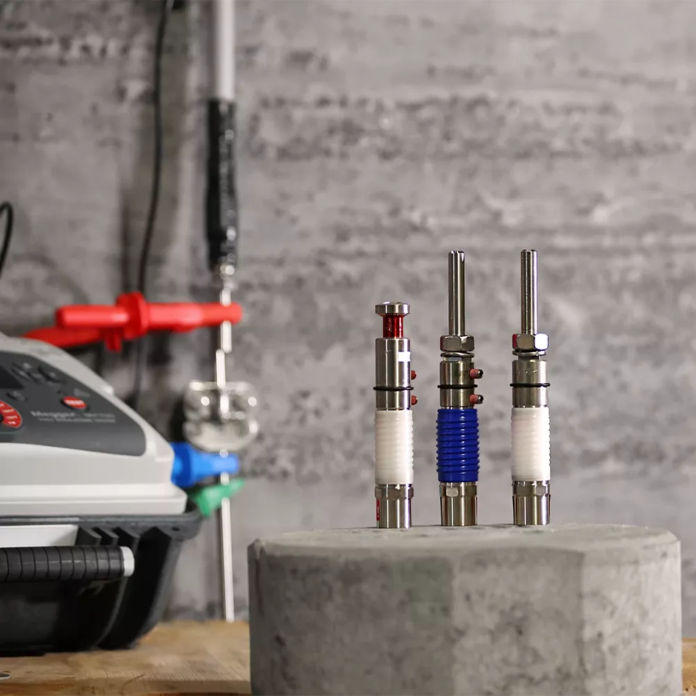 Tres objetos cilíndricos de metal con componentes rojos, azules y blancos sobre una superficie de hormigón delante de un equipo con un panel de control.